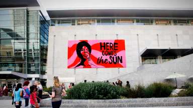 Large scale crocheted portrait of Nina Simone with the words &quot;Here Comes the Sun&quot; hanging on the outside of the Raleigh Convention Center.
