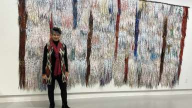 Artist, a slight, white woman with short grey hard, in front of a textured fiber wall hanging in a museum.