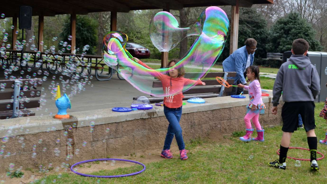 Young girl blows a large oblong bubble in an arc near short stone wall at Anderson Point Park
