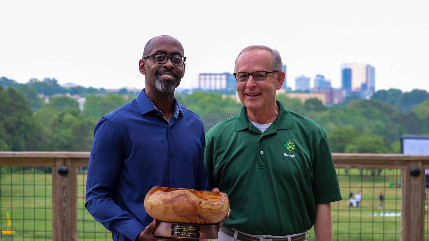 Two members of Walnut Creek Watershed Team pose with award with Raleigh skyline in background