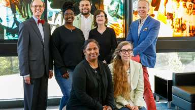 Portrait of seven people who serve on the arts commission. They are lined up and smiling for the camera in front of an art glass window.