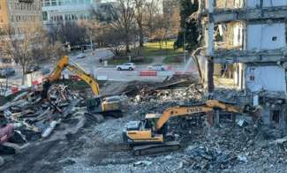 Construction debris sorting by backhoe and large claw machine, also in the image is the shell of the old police HQ
